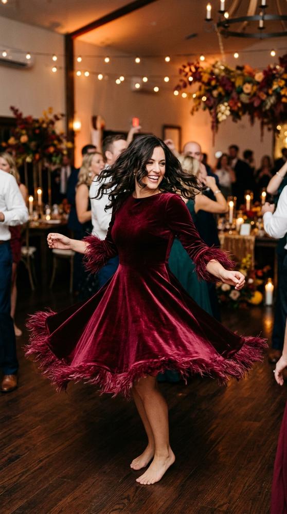 Woman in a burgundy feather-trim cocktail dress twirling on a dance floor at a fall wedding