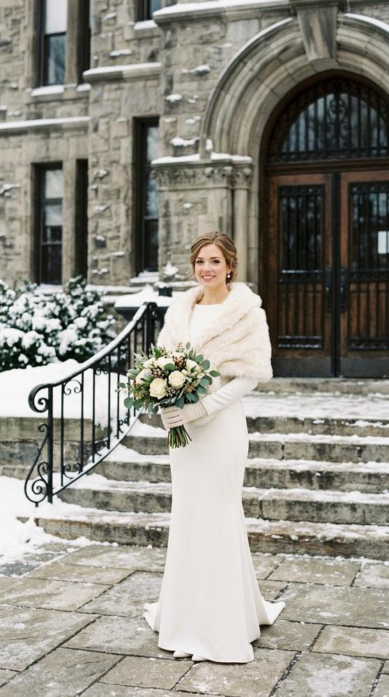 A bride in an ivory long-sleeve crepe wedding dress with a faux fur stole outside a snow-dusted courthouse