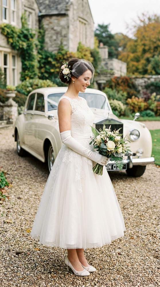A bride wearing elbow-length ivory wedding gloves with a tea-length dress, vintage car behind.