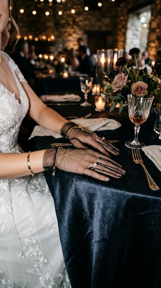 Bride wearing mesh gloves with delicate chain details connecting the fingers, hands over a dark silk tablecloth.