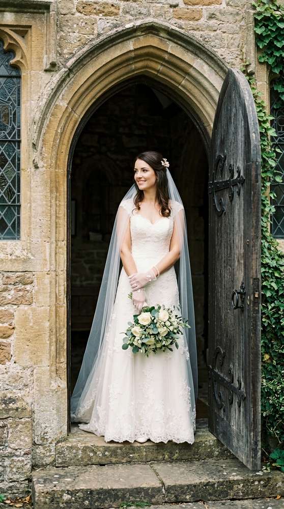 Bride in classic white mesh wrist gloves with a timeless A-line gown, standing in a historic church doorway.